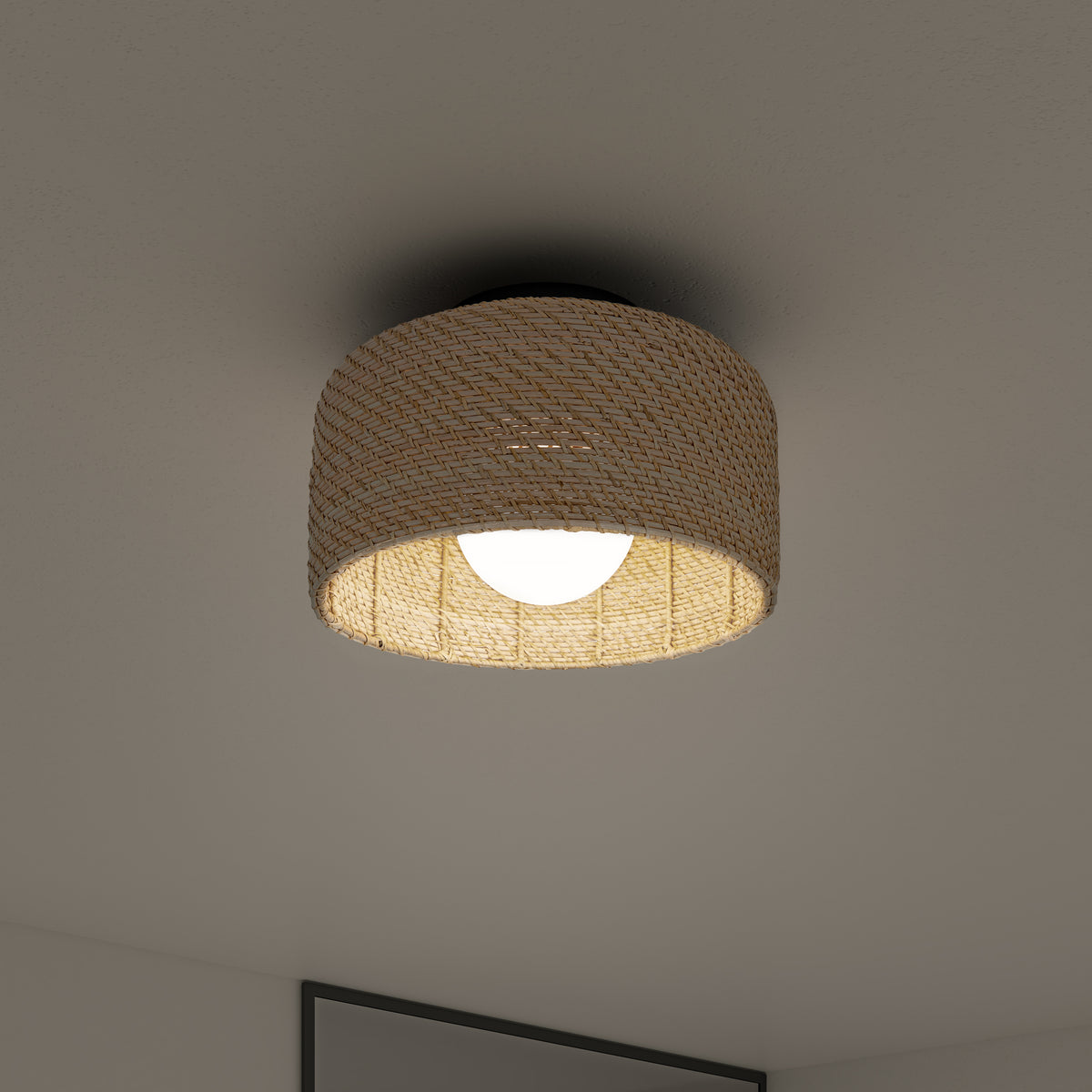 A round ceiling light with a woven, beige lampshade emits a soft glow, mounted on a smooth white ceiling above a partially visible gray-framed picture.