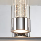 A close-up of a modern light fixture with a polished chrome base and a cylindrical glass shade featuring small bubbles inside, mounted on a reflective surface. The light inside is turned on.