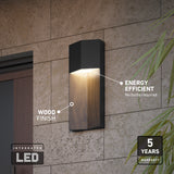 A modern black and wood-finish wall light is mounted on a stone wall, labeled as energy efficient with integrated LED and a 5-year warranty. Green plant leaves are visible on the right side.