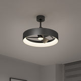 A modern black ceiling fan with a circular LED light fixture is mounted on a white ceiling in a minimalistic room with gray walls and a window with gray blinds.