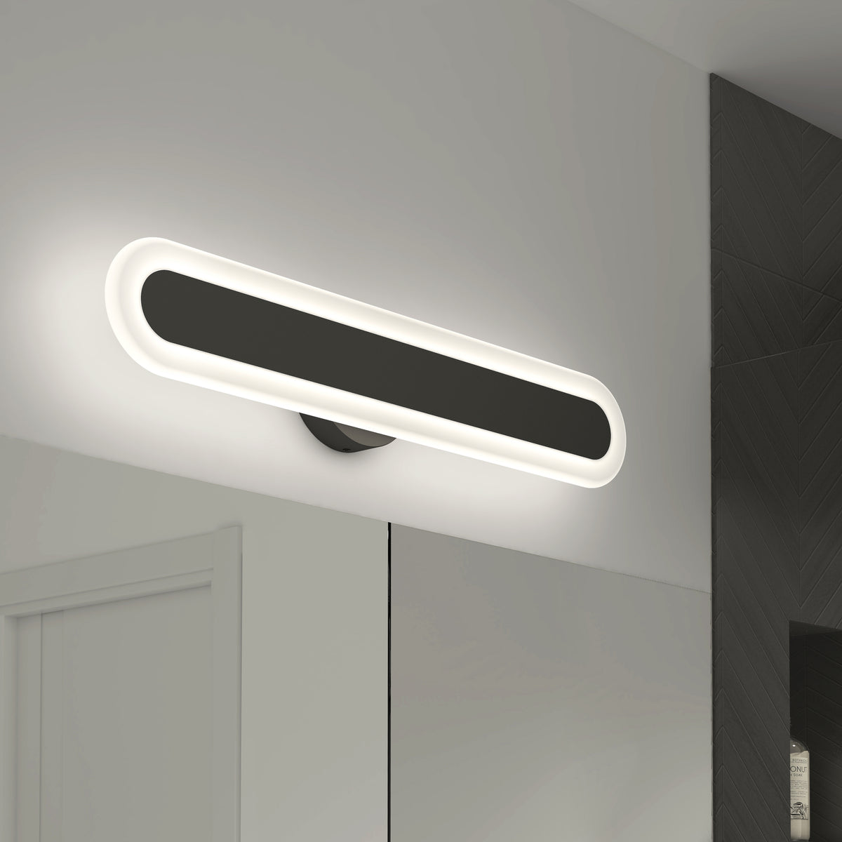 A modern, oval-shaped wall light fixture with a glowing white border mounted above a mirror in a minimalist bathroom with white and dark gray walls.