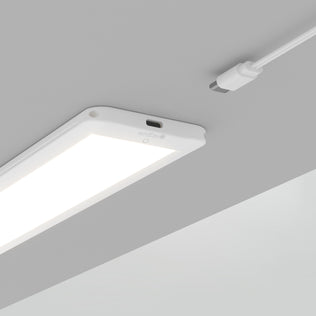 The Artika Maestro under cabinet LED light 3CCT is illuminated on a gray ceiling, featuring a white rectangular design with a white power cable and connector hanging nearby.