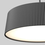 Close-up of a modern, cylindrical pendant lamp with a black, vertically ribbed lampshade, glowing warmly from inside, set against a plain light gray background.