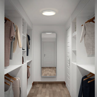 A modern walk-in closet with white shelves, hanging clothes, a mirror, wood flooring, and an Artika Vera 13 in. LED ceiling light.
