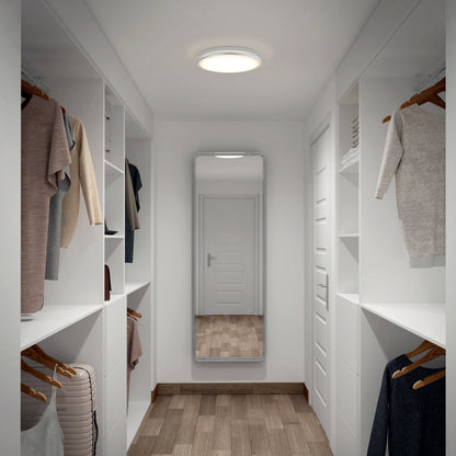 A modern walk-in closet with white shelves, hanging clothes, a wooden floor, a closed white door reflected in a full-length mirror, and an Artika Vera 13 in. LED modern flush mount ceiling light providing bright, dimmable illumination.