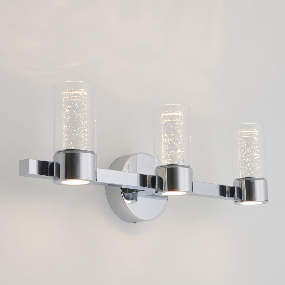 A modern wall sconce with three cylindrical glass light fixtures featuring a sparkling, bubble-like design, all mounted on a chrome base against a white wall.