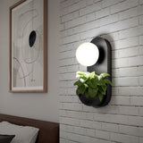 A modern wall sconce with a round white light and a built-in planter holding green leaves, mounted on a white brick wall beside abstract art and a brown bed headboard.