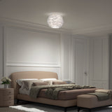 A neatly arranged modern bedroom with a beige upholstered bed, green bedding, matching ottoman, nightstand with flowers, and a white feathered ceiling light illuminating the space.