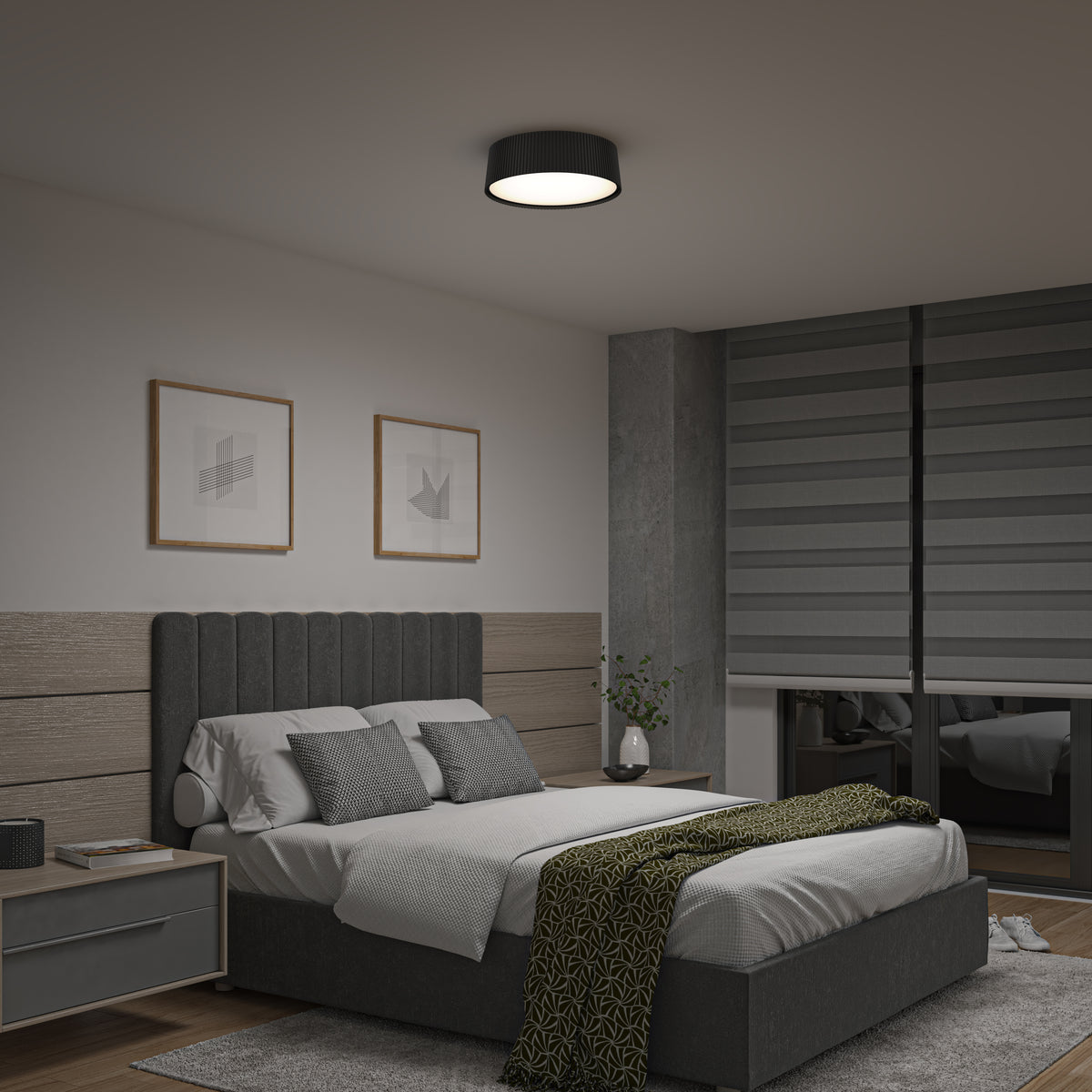 A modern bedroom with a gray upholstered bed, green patterned throw, two framed abstract artworks above the headboard, a nightstand with a lamp, and large gray blinds covering the windows.