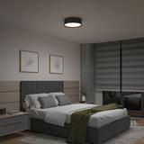 A modern bedroom with a gray upholstered bed, green patterned throw, two framed abstract artworks above the headboard, a nightstand with a lamp, and large gray blinds covering the windows.