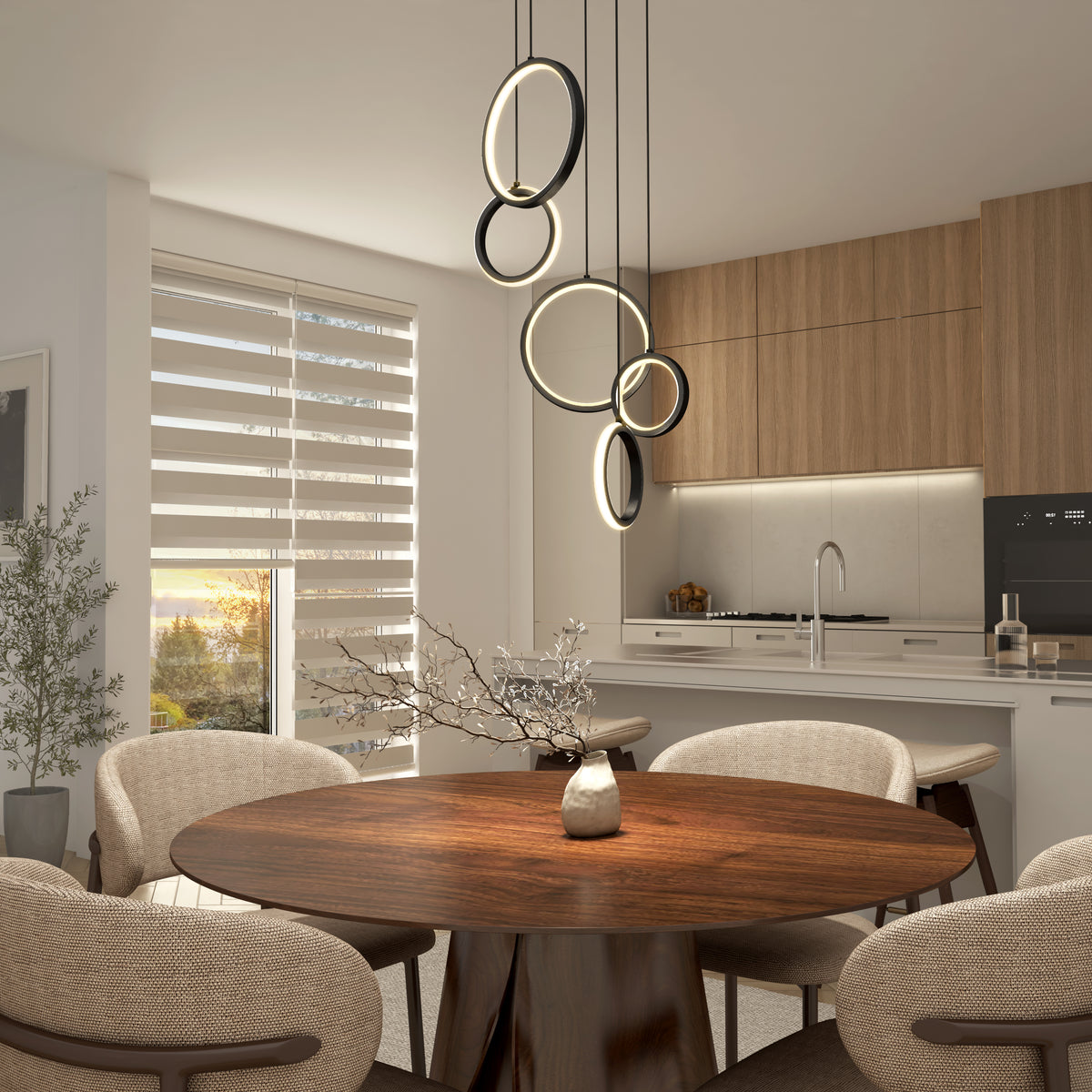 A modern dining area with a round wooden table, beige upholstered chairs, a decorative branch centerpiece, stylish ring pendant lights, and a kitchen with wood cabinets in the background. Sunlight streams through layered blinds.