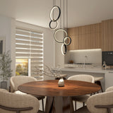 A modern dining area with a round wooden table, beige upholstered chairs, a decorative branch centerpiece, stylish ring pendant lights, and a kitchen with wood cabinets in the background. Sunlight streams through layered blinds.