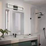 A modern bathroom with a large mirror, rectangular wall light above it, double sink with black faucets, green vanity, glass shower enclosure, and minimal decor including a small plant and bottle.