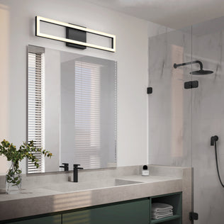 A modern bathroom with a large mirror, an Artika Dover 27-in Integrated LED vanity light above, double sinks with black faucets, a glass shower enclosure, black shower fixtures, and a vase with green foliage on the counter.