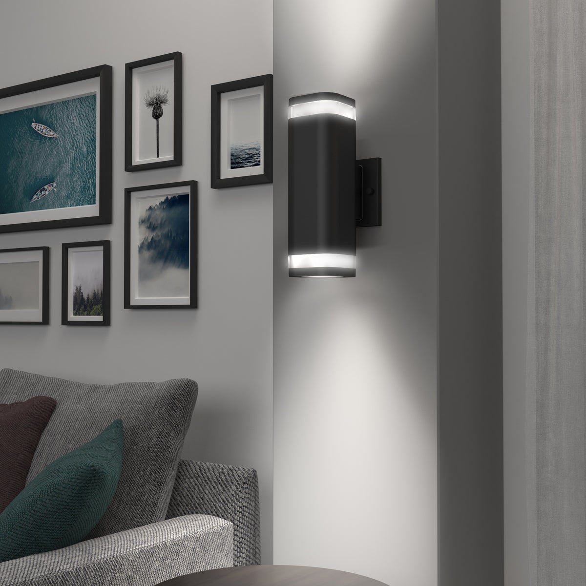 A modern black wall sconce with upper and lower LED lights is mounted on a white wall near a gray sofa. Framed photos and artwork are arranged on the adjacent wall.