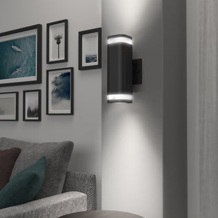 The Artika Stark modern LED indoor-outdoor light with Wi-Fi is mounted on a light gray wall above a gray sofa, next to a gallery of framed photos and artwork.