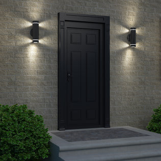 A black front door with decorative panels sits in a beige brick wall, flanked by two Artika C7 indoor-outdoor wall lights. Two small green bushes frame the entry, and a stone step leads up to the door.