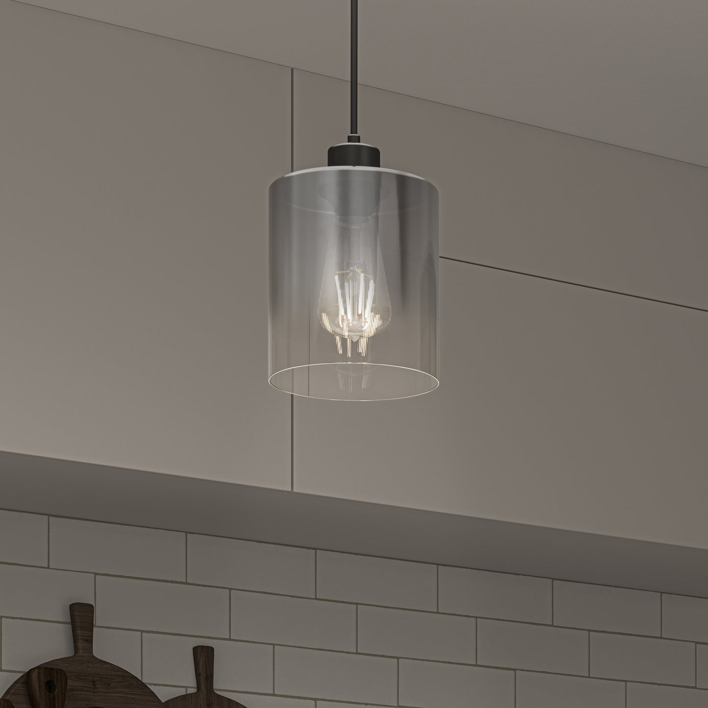 The Artika Haze pendant light with a clear cylindrical glass shade hangs in a kitchen featuring white subway tile backsplash, beige cabinets, and two wooden cutting boards on the counter below.