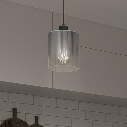 The Artika Haze pendant light with a clear cylindrical glass shade hangs in a kitchen featuring white subway tile backsplash, beige cabinets, and two wooden cutting boards on the counter below.