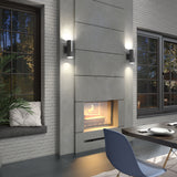 Modern living room with a concrete fireplace and two wall sconces above it, a window seat with cushions and a plant, large windows, and a dining table with a blue chair in the foreground.