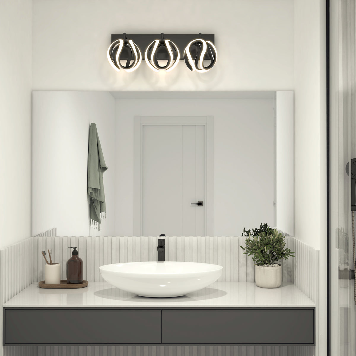 Modern bathroom with a round white sink, black faucet, potted plant, soap dispensers, a towel on the wall, and a large mirror reflecting a white door. Above the mirror is a contemporary three-light fixture.