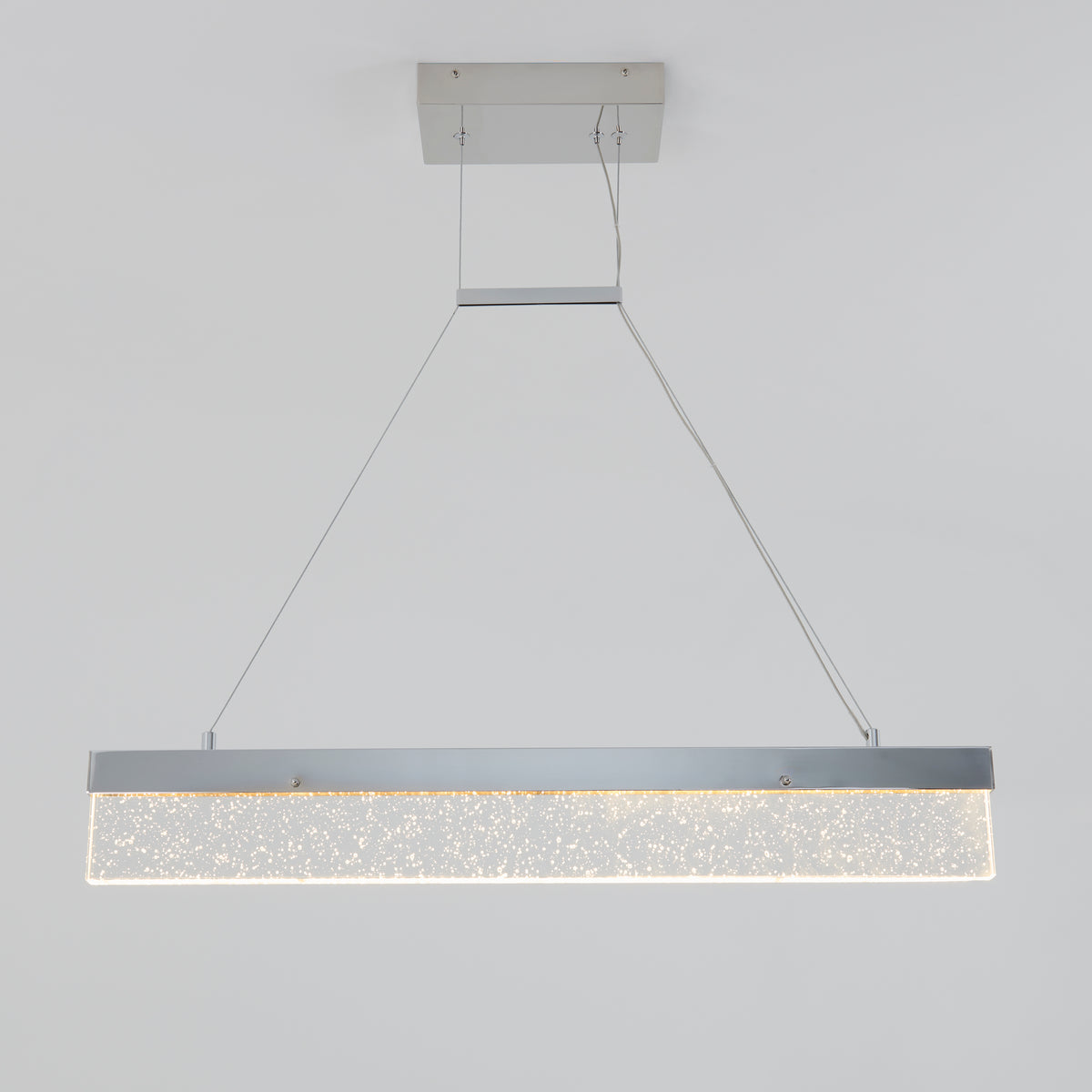 A modern rectangular pendant light fixture with a metallic frame and sparkling bubble-like details hangs from the ceiling by two thin cables. The light is illuminated against a plain white background.