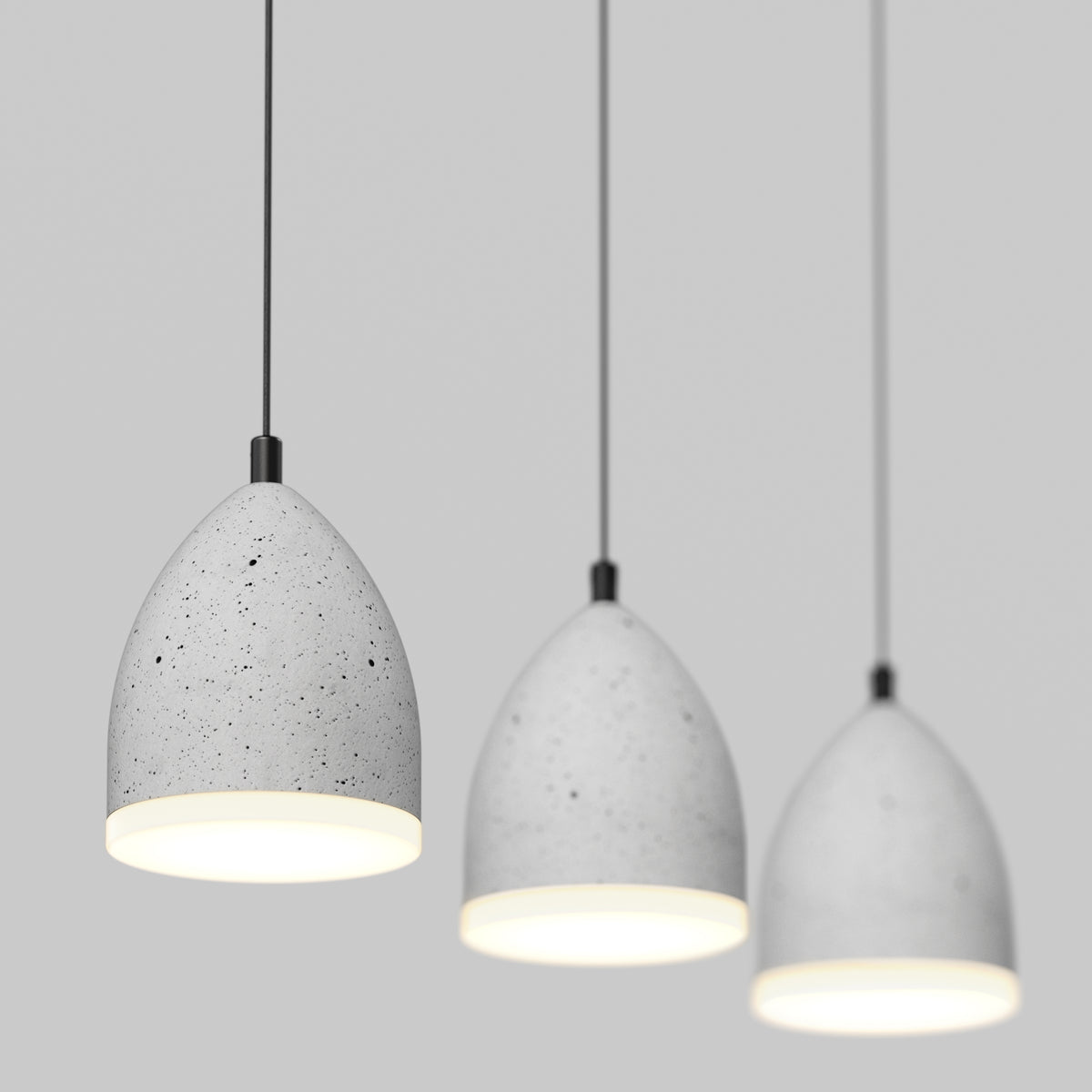 Three modern pendant lights with smooth, speckled gray concrete shades hang from black cords against a plain light gray background. The lights are lit, casting a soft white glow downward.