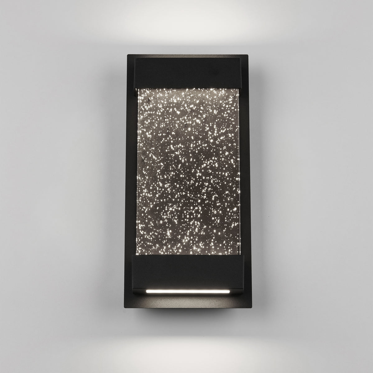 The Artika Essence Glow Box indoor-outdoor LED wall light features a modern rectangular design with a black frame and star-textured illuminated glass, casting soft up-and-down lighting against a light gray wall.