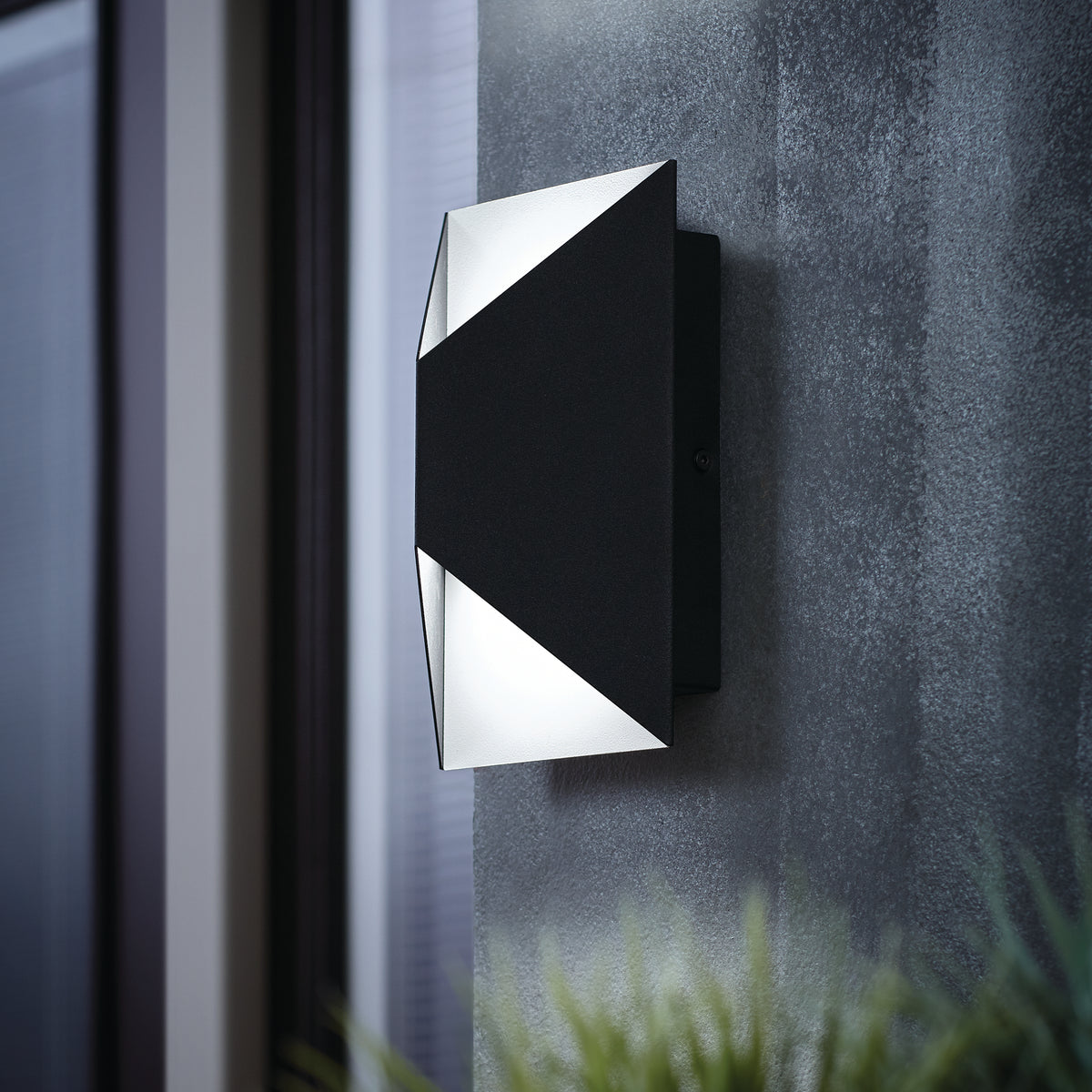 A modern black and white wall sconce with geometric design is mounted on a textured gray wall, emitting soft light. A window and some blurred indoor plants are visible in the background.