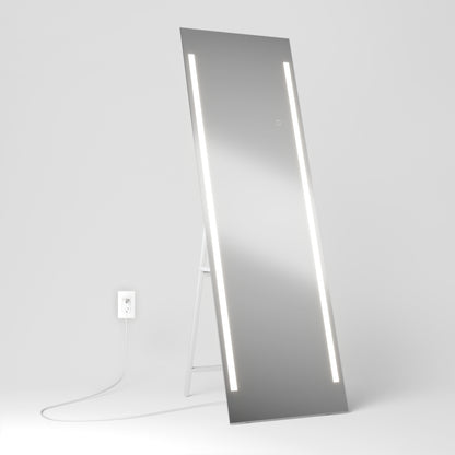 The Artika Emeraude Full Length Mirror features a tall rectangular design with dual vertical LED strips on each side, leaning against a white wall and plugged into a nearby outlet. The minimal, bright background highlights its modern style.
