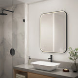 A modern bathroom with a large, illuminated rectangular mirror above a white vessel sink, black fixtures, soap dispenser, and a glass-enclosed shower with marble walls. A small plant sits on the countertop.