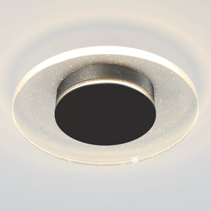 The Artika Essence Disk LED flush mount is a modern ceiling light featuring a black center, transparent outer ring with sparkling specks, and emits a soft white glow.