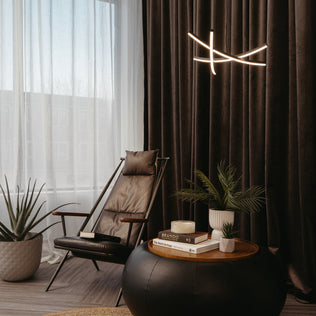 A modern lounge with a black leather chair, potted plants, stacked books, and a candle on a round ottoman. Sheer curtains cover large windows, while the Artika Orly modern LED pendant light 5 CCT hangs above.