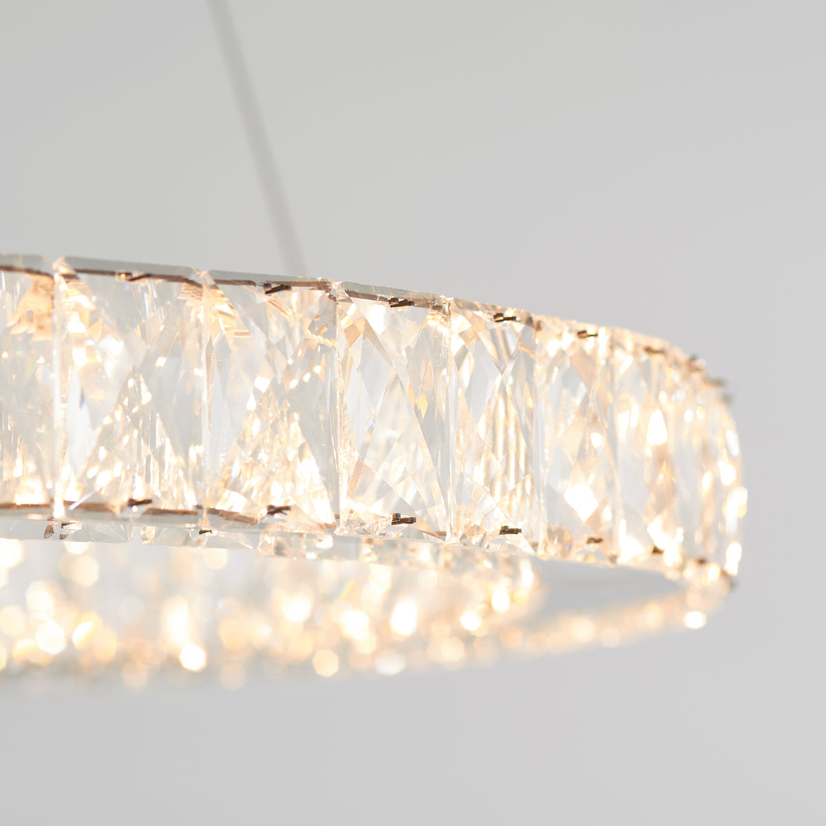 Close-up of a circular crystal chandelier with faceted glass panels, emitting a warm, soft light against a plain light gray background.