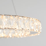 Close-up of a circular crystal chandelier with faceted glass panels, emitting a warm, soft light against a plain light gray background.