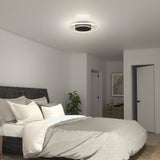 A modern bedroom with a gray upholstered bed, white and gray bedding, a bedside plant, a wooden dresser, and a round ceiling light fixture. Walls are white and the room has a minimal, clean decor style.