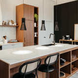 Modern kitchen with a large marble island, two black chairs, pendant lights, wood cabinets, open shelves with decor, and a black accent wall. A sink is built into the island and plants add a touch of greenery.