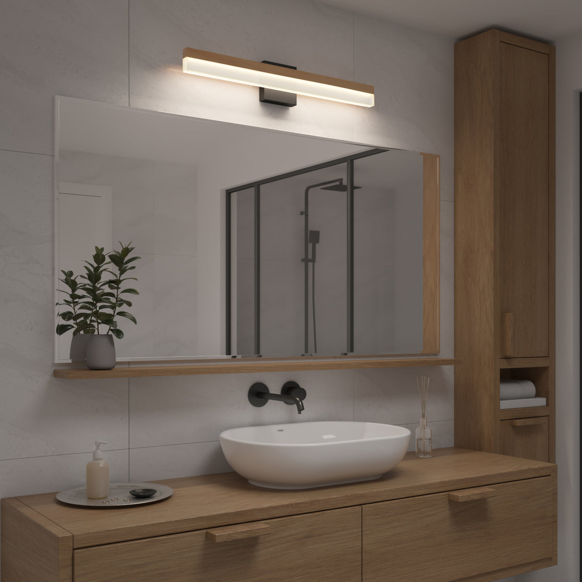 A modern bathroom with a wooden vanity, a white vessel sink, a large mirror, a wall-mounted light fixture above the mirror, a potted plant, soap dispenser, and rolled towels on a shelf. A shower is visible in the background.