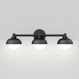 A modern wall-mounted light fixture with three round bulbs, each covered by a matte black dome-shaped shade, set against a plain light gray background.