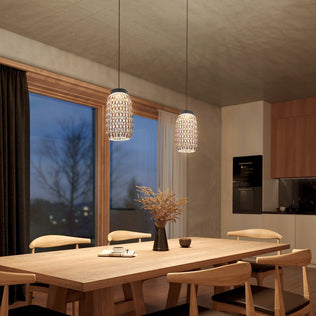 Modern dining room featuring a wooden table and chairs, Benson Mini LED pendant lights by Artika hanging above, a vase with dried branches, large window with dark curtains, and a kitchen area in the background.