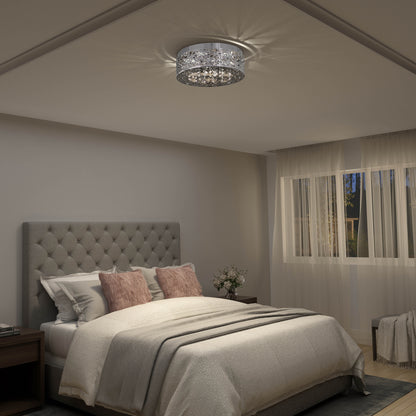 A cozy bedroom features a tufted gray headboard, white bedding, blush pink pillows, and Artika’s Crystal Nest LED flush mount. Sheer curtains cover large windows, and a bedside table holds a small flower vase.
