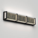 A modern wall-mounted light fixture with three rectangular panels featuring textured, sparkling glass and a sleek black metal frame, emitting a soft white glow against a light gray wall.