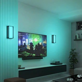 A modern living room with Artika’s Ryker smart RGB indoor-outdoor wall light in teal, a wall-mounted TV showing a sci-fi scene, floating shelves, a game console, and a potted plant in the corner.