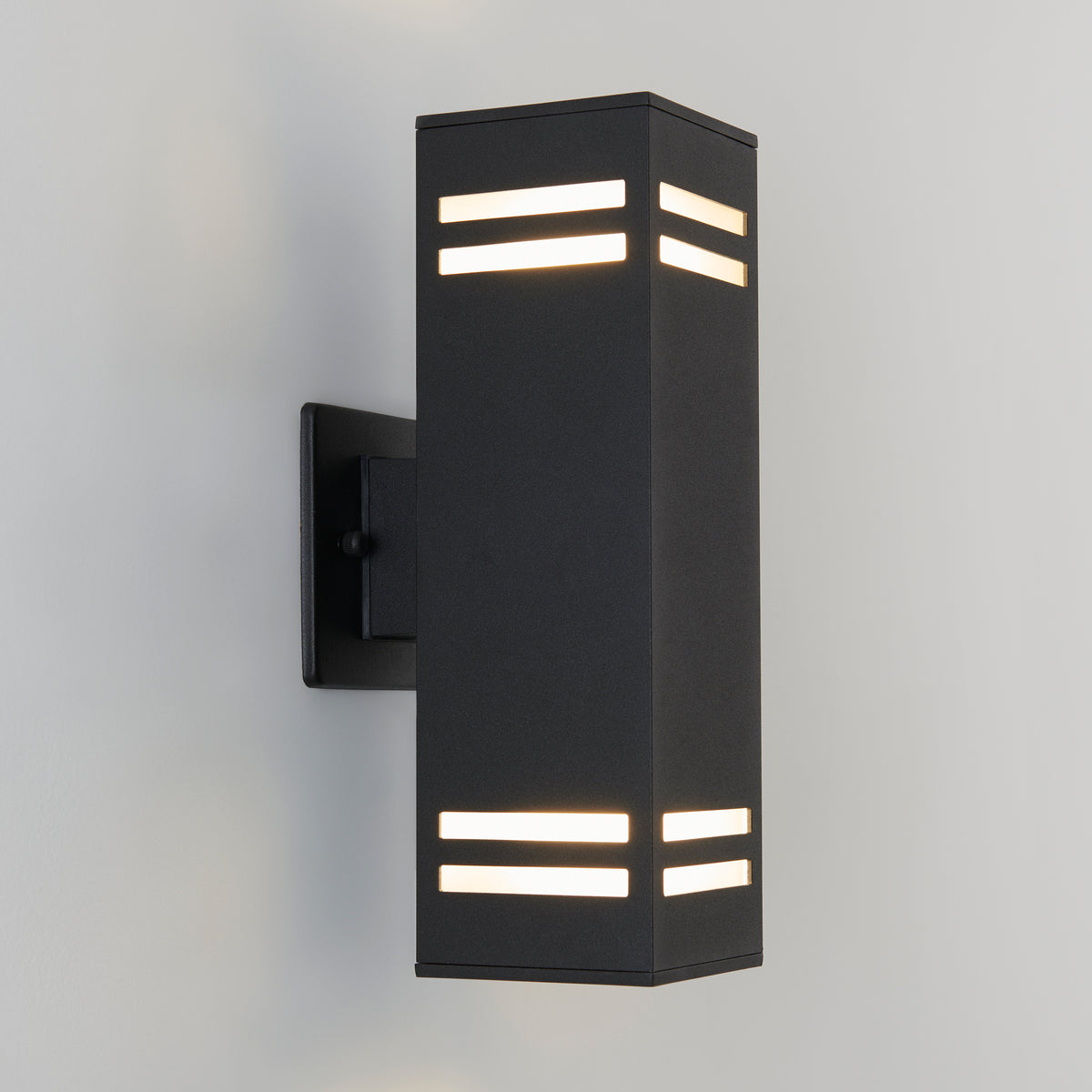 The Artika Strike indoor-outdoor wall light is a modern black rectangular sconce featuring horizontal top and bottom slits, mounted on a light-colored wall and emitting soft white light.