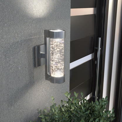 The Artika Bubble Element LED indoor-outdoor light, featuring a metallic frame and bubble-patterned glass, is mounted on a gray wall near a door above green leafy plants, casting an inviting glow.