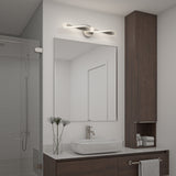 A modern bathroom with a rectangular mirror above a sink, a sleek looped light fixture above the mirror, a wooden cabinet, glass shower, and neatly arranged towels and plants on the counter.