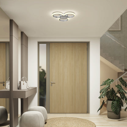 A modern entryway features a wooden door, mirror, plant, beige poufs, woven rug, handbag on a narrow table, and the Artika Nyala 15 in. LED flush mount light 5CCT overhead. A staircase with a glass railing appears on the right.