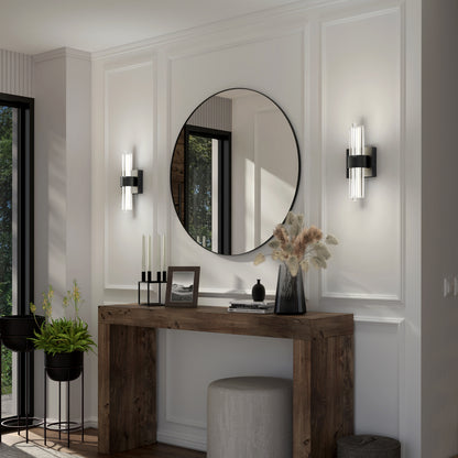 A modern entryway featuring a rustic wooden console table, round mirror, Artika Dorado LED contemporary wall sconces, decorative vases, candles, potted plants, and a round upholstered stool set against white paneled walls.
