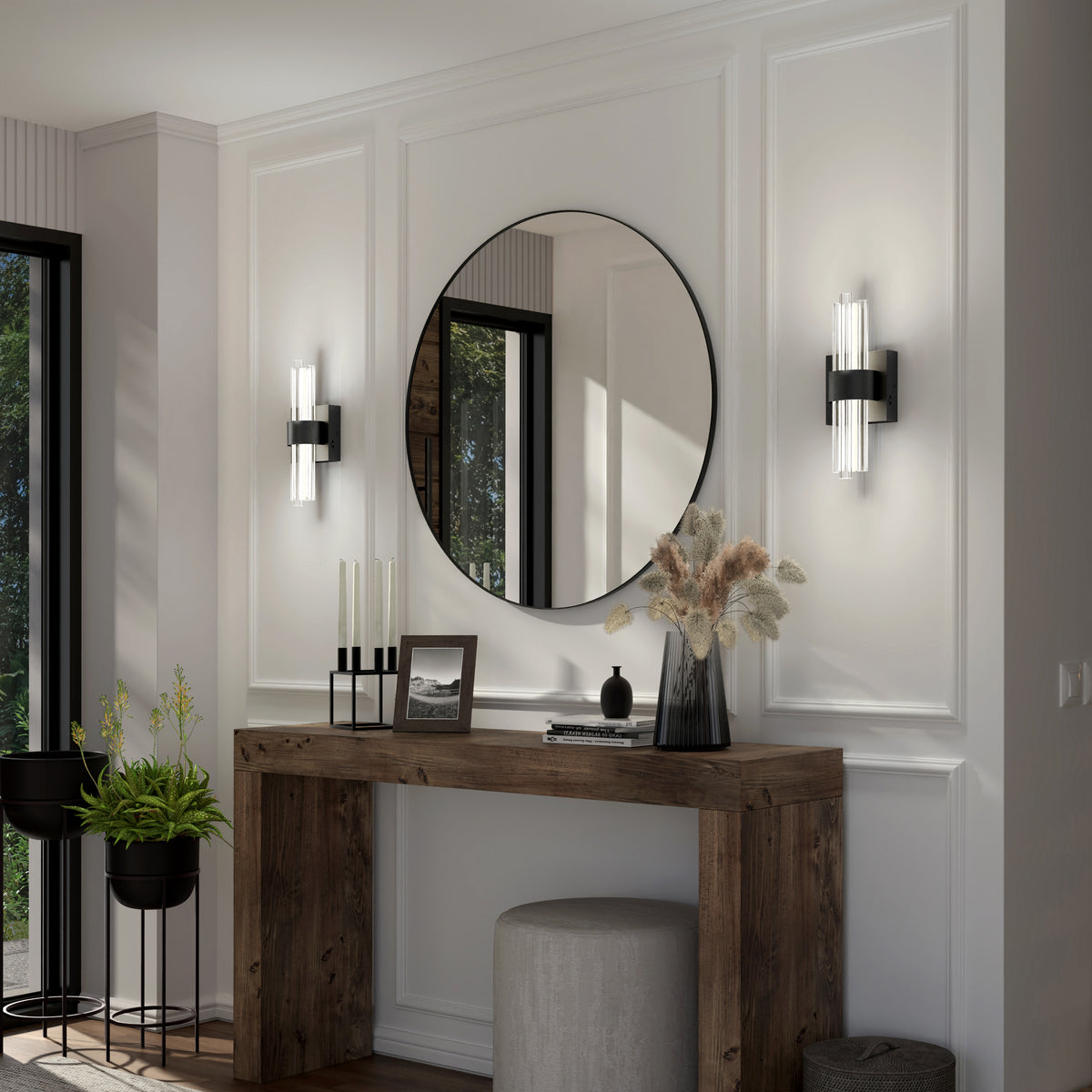 A modern entryway with a rustic wooden console table, stool, decorative vase with dried flowers, candles, round mirror, wall sconces, and potted plants against white paneled walls.