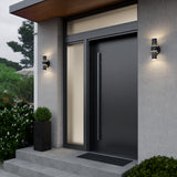 Modern front door with a sleek black finish, large vertical handle, side and transom windows, two wall-mounted lights, a square planter with greenery, and concrete steps leading up to the entrance.